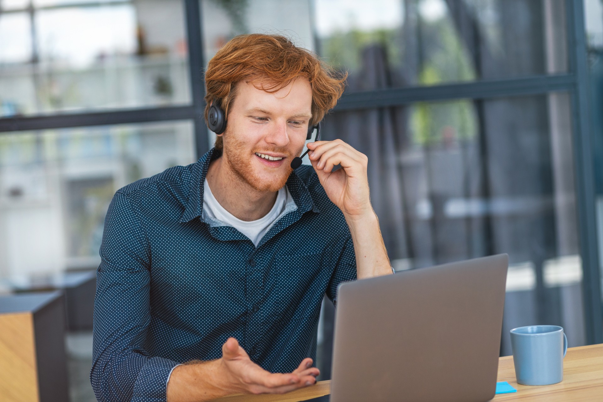 Kaukasischer junger Mann Kundensupport-Call-Center-Operator oder Rezeptionist, der am Arbeitsplatz in einem modernen Büro sitzt und einen Kunden berät, ein Headset verwendet, freundlich lächelt