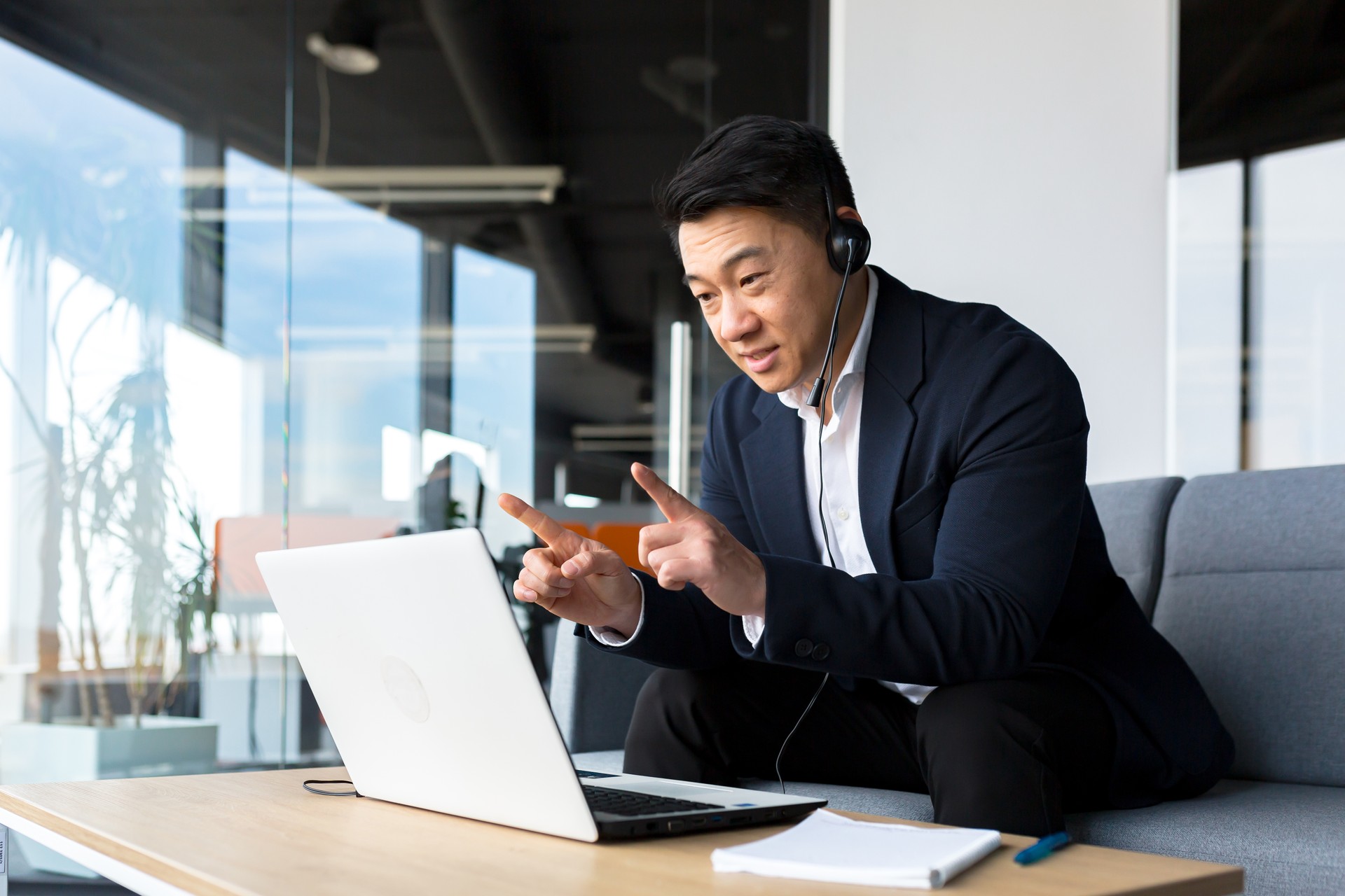 Asiatischer Call-Center-Mitarbeiter im Gespräch mit Kunden, die im Büro sitzen, asiatischer Geschäftsmann mit Headset und Laptop für Videoanruf glücklich und lächelnd