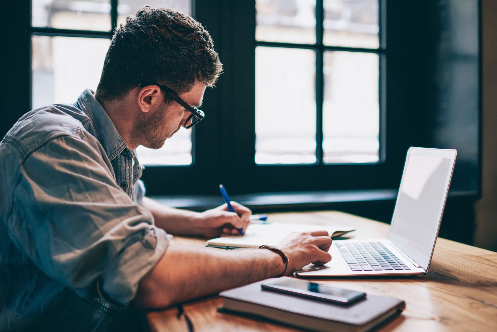 Junge Millennial männlichen Studenten auf der Suche nach Informationen für Hausaufgaben mit Laptop und WiFi-Verbindung in Innenräumen, Mann schreiben Artikel in Lehrbuch für interessante Inhalte auf eigener Website oder Webseite hinzufügen